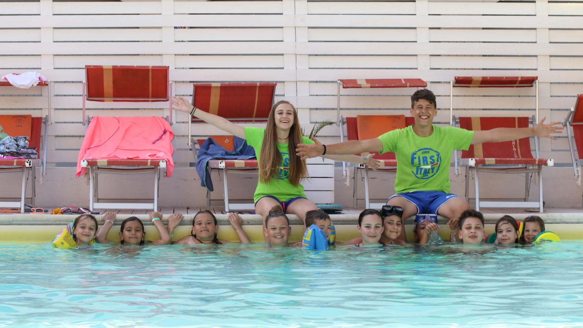 Gruppo di bambini in piscina con due istruttori sorridenti in maglietta verde.