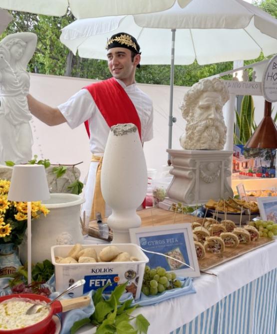 Stand greco con cibo tradizionale e decorazioni classiche, uomo in costume storico.