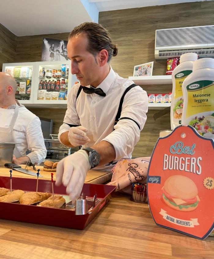 Uomo prepara panini in una cucina, cartello 'Best Burgers' in vista.