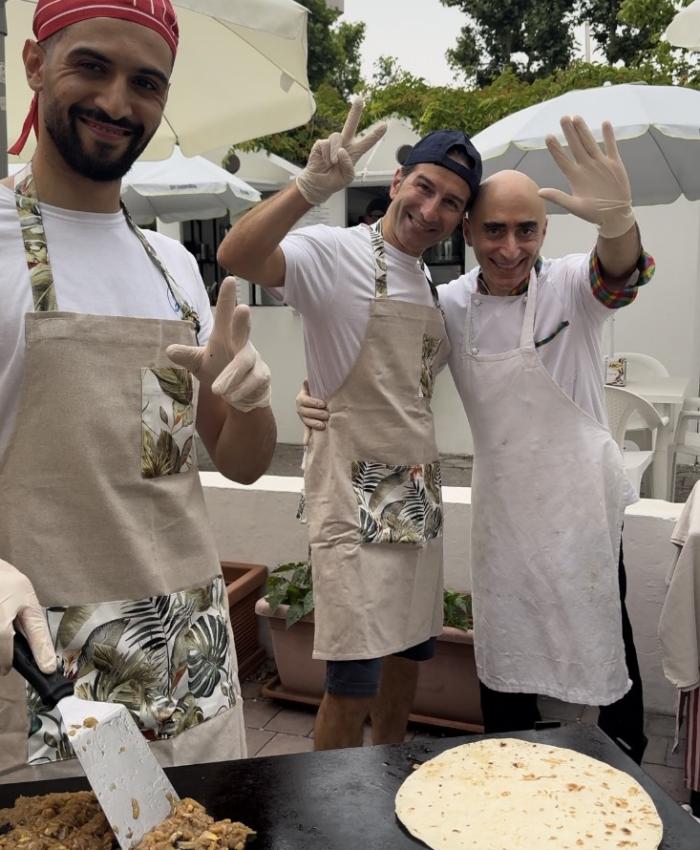 Tre cuochi sorridenti con grembiuli, cucinano all'aperto con piadine su una piastra.